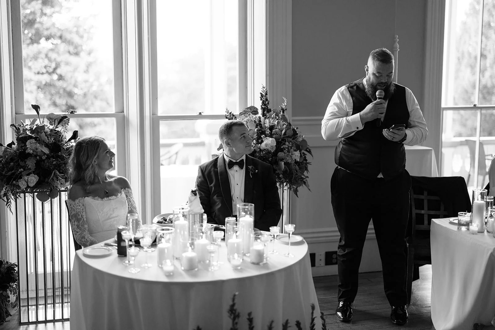 A black and white photo of a wedding reception with a man giving a speech, standing by a table with candles, and the bride and groom sitting nearby, smiling and listening.
