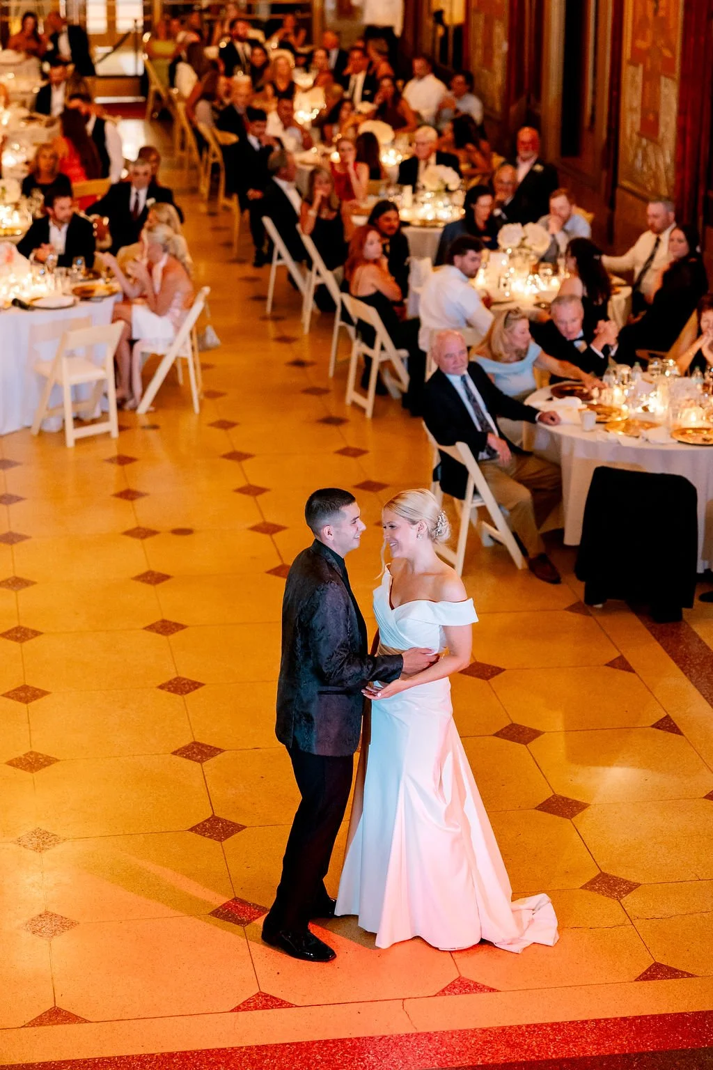 A bride and groom dance during their wedding reception in a decorated banquet hall filled with seated guests.