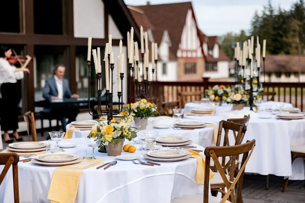 Elegant outdoor dining setup with white tablecloths, floral centerpieces, candles, and place settings, with musicians playing violin and keyboard in the background.