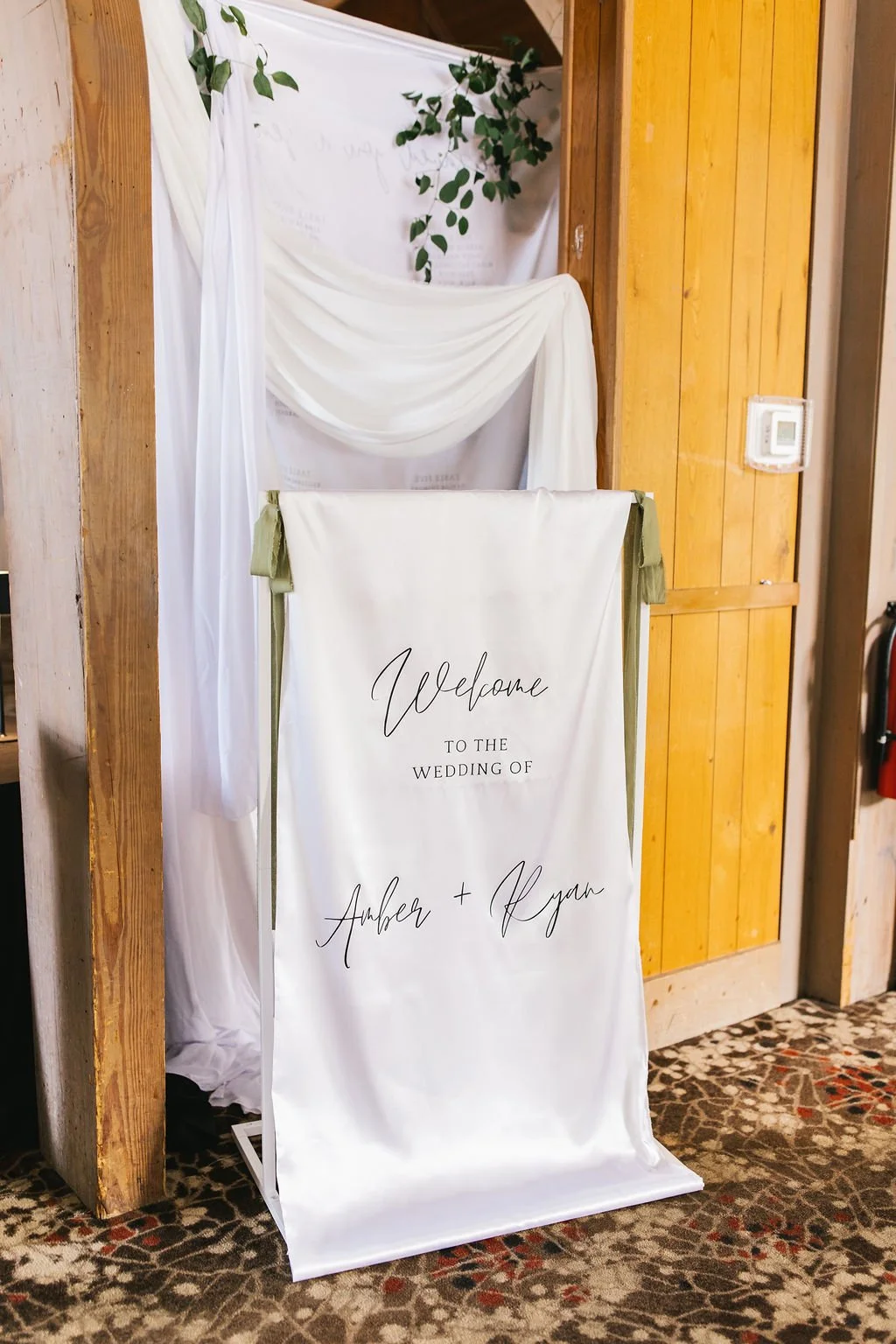 A wedding welcome sign for Amber and Aegan, placed on an easel, with decorative drapery and greenery in the background.