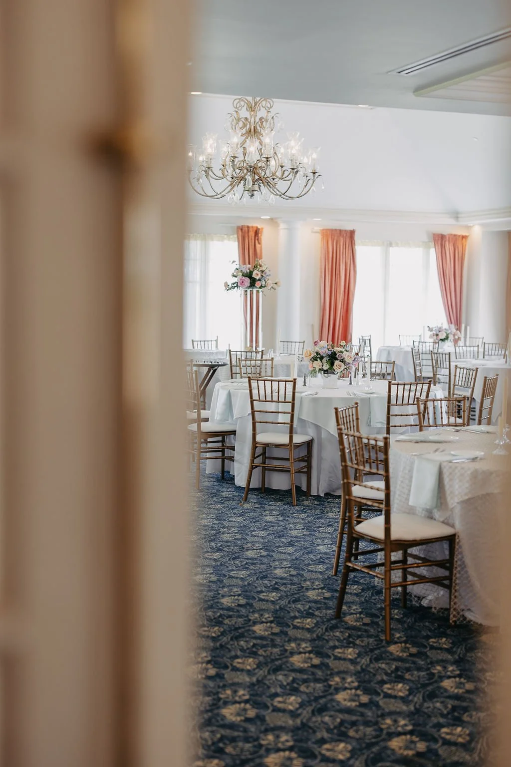 Elegant banquet hall with round tables covered in white tablecloths, decorated with pink and white flower centerpieces, surrounded by gold chairs. A chandelier hangs from the ceiling, and tall windows with pink curtains allow natural light to fill th