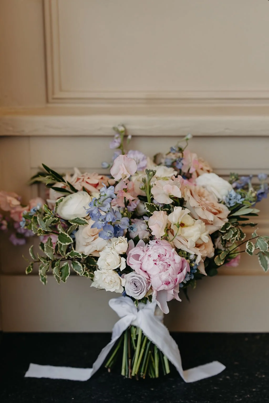 A bouquet of pastel-colored flowers including roses, peonies, and delphiniums, tied with a white ribbon, resting against a beige wall.