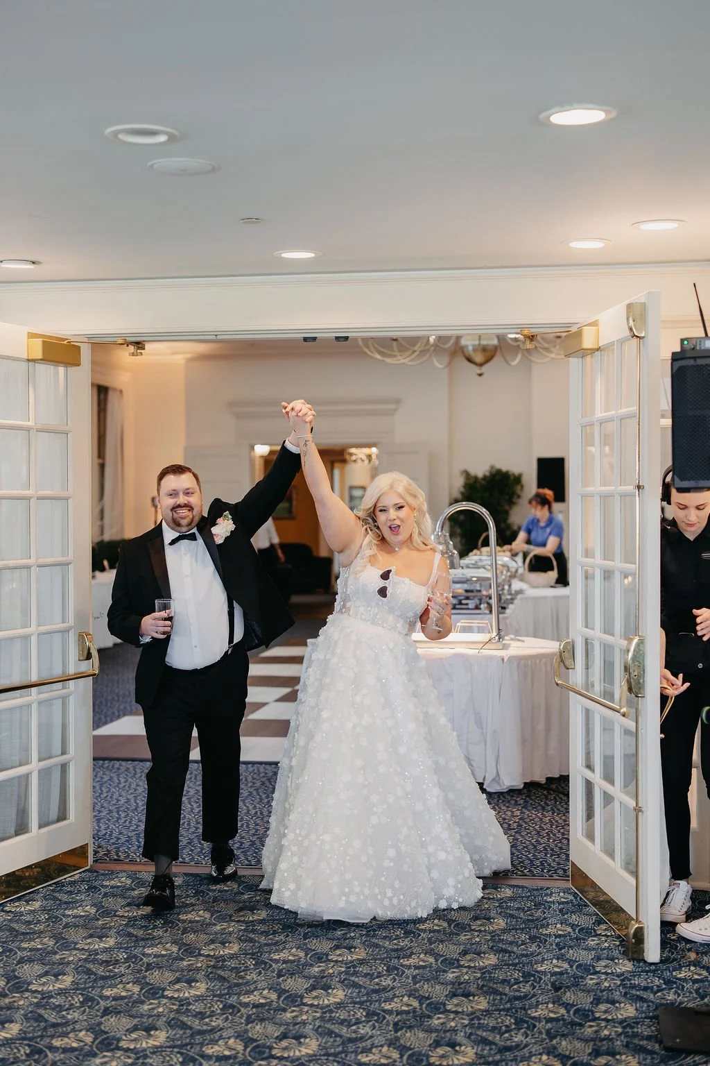 Bride and groom celebrating at their wedding reception as they exit through open gates, with heightening joy. The bride has blonde hair and is wearing a white wedding gown, and the groom has a beard and is wearing a black tuxedo. They are holding han