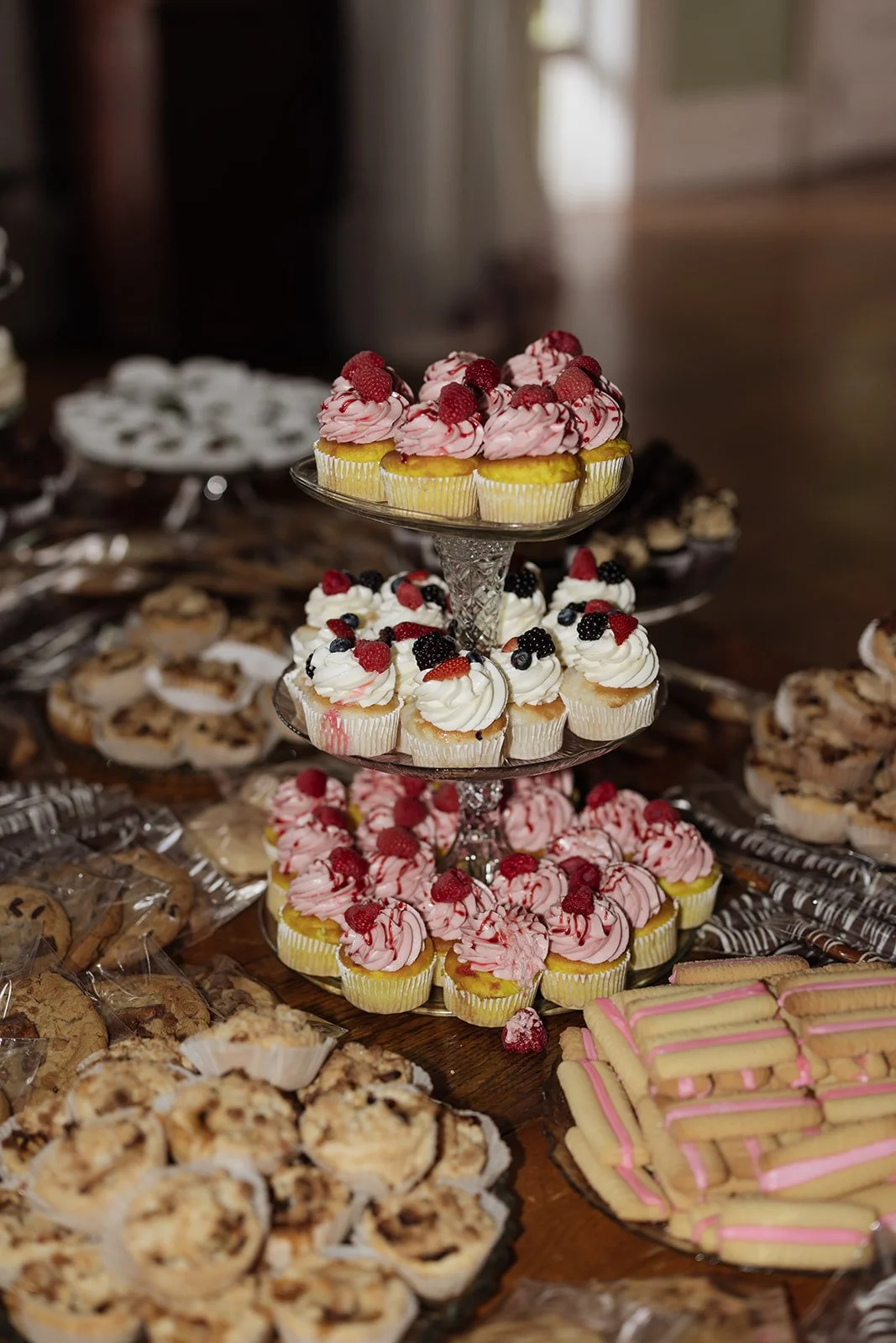 Assorted cupcakes with pink and white frosting topped with raspberries, blueberries, and blackberries on a multi-tiered glass stand, surrounded by various cookies and pastries on a wooden table.