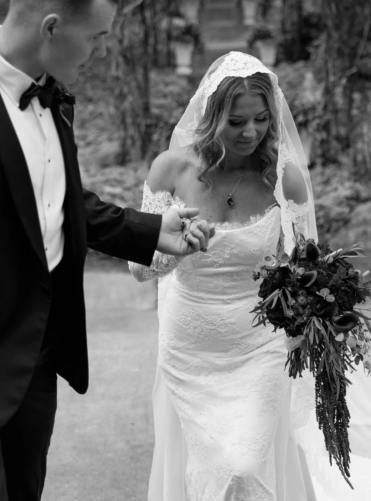 A bride with a lace off-the-shoulder wedding dress holding a bouquet of flowers, and a groom in a tuxedo, holding hands during a wedding ceremony outside.