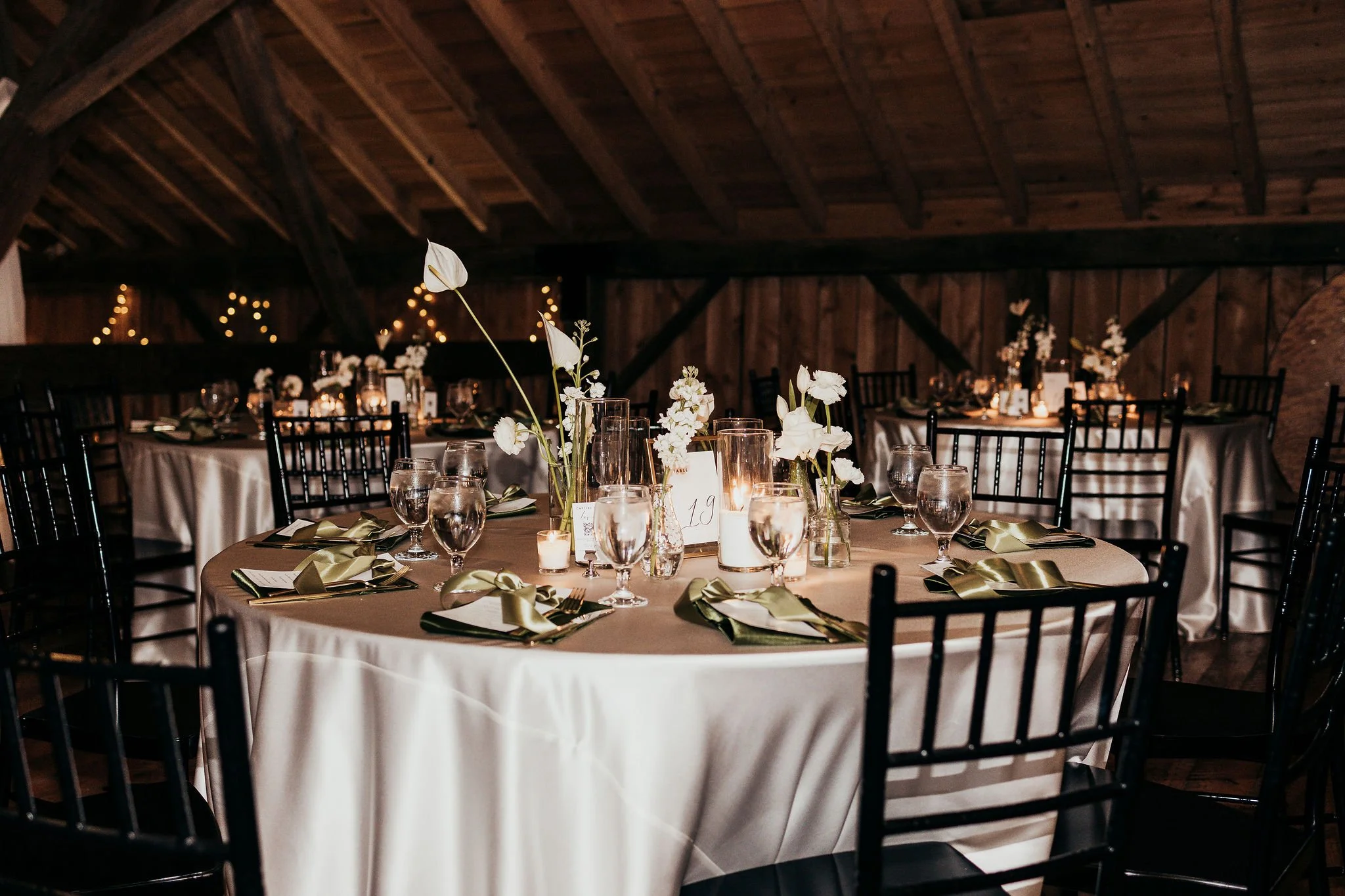 Elegant round table set for a formal event with white tablecloth, clear glassware, cutlery, and white floral centerpieces, decorated with gold ribbons and surrounded by black chairs in a rustic wooden venue with soft lighting.
