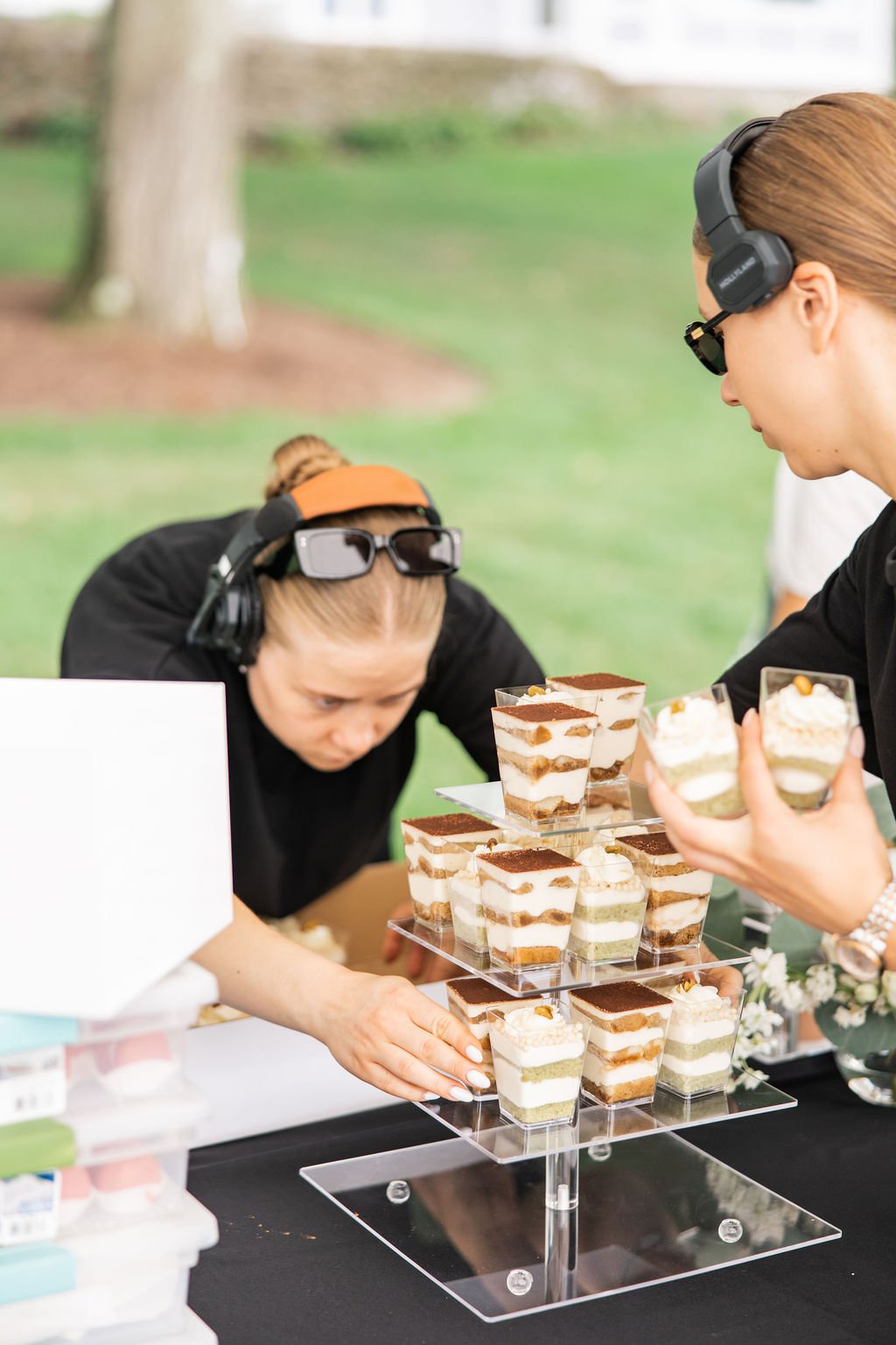 Two women wearing headphones and sunglasses are at a dessert stand, one of them is arranging small layered cakes on a clear acrylic display stand.