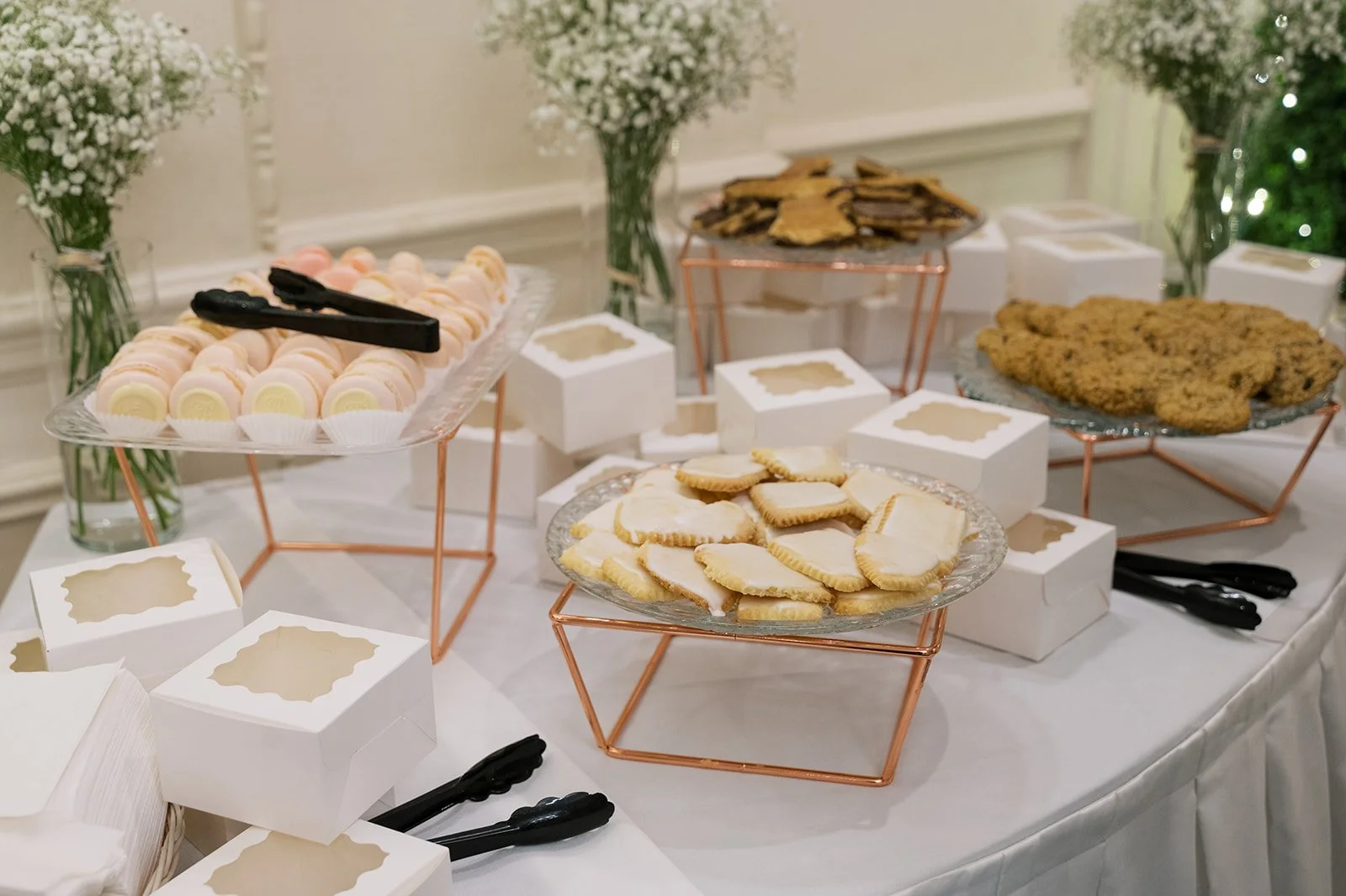 Dessert table with pink and white macarons, sugar cookies, chocolate chip cookies, and chocolate bark on elevated trays. White boxes with window cutouts surround the desserts, with white flowers in vases in the background.