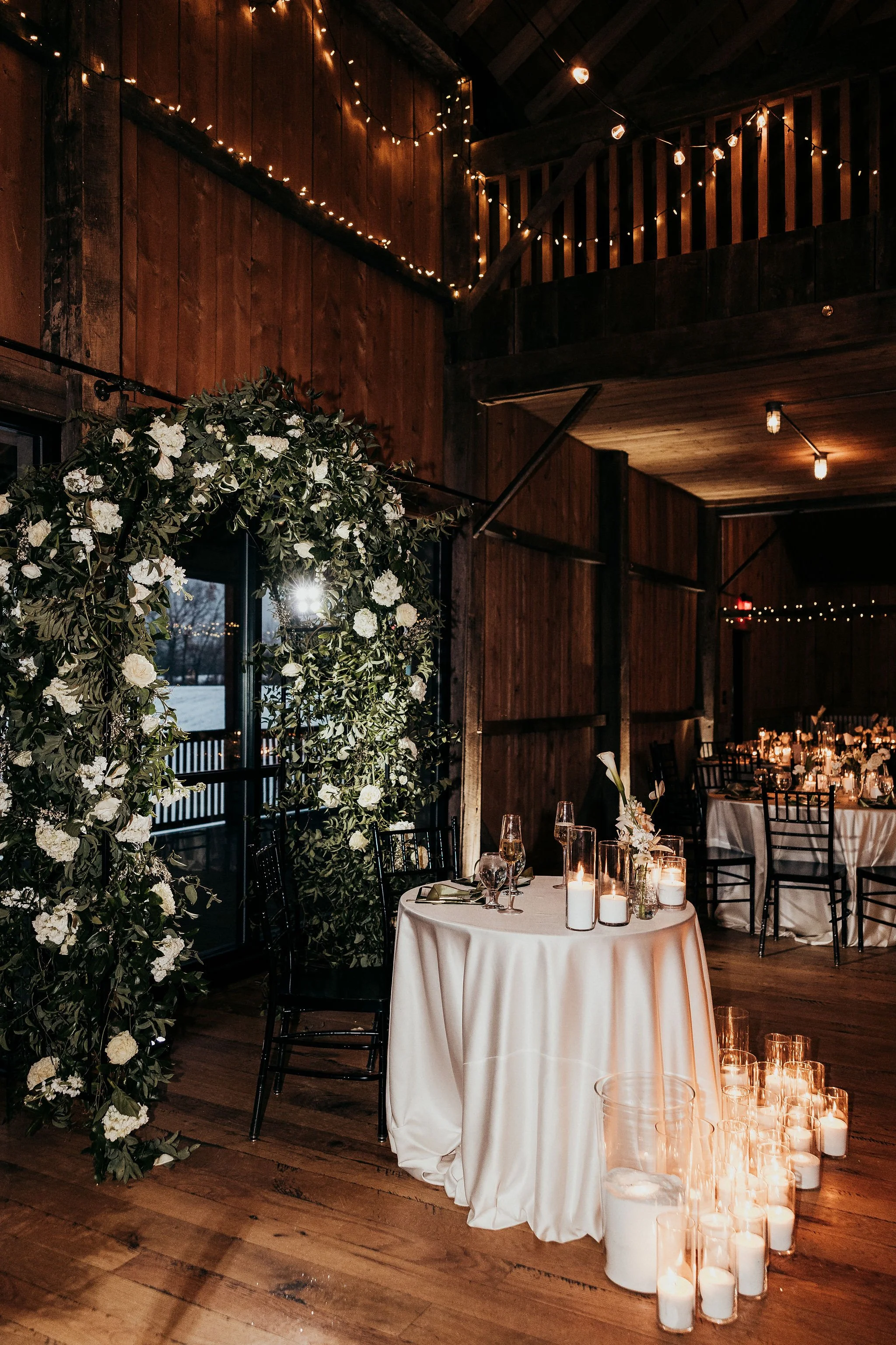A decorated indoor event space with wooden walls and ceiling, string lights, a large floral arch, and a table with candles and glassware for a celebration, possibly a wedding reception.