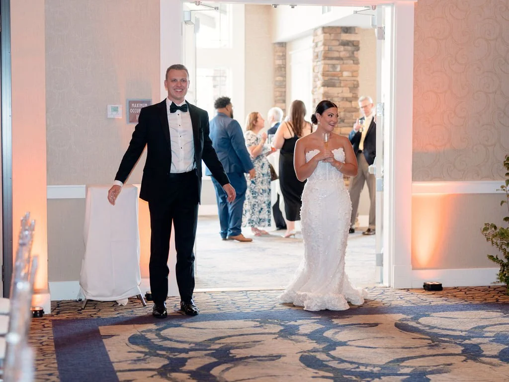 Bride in a white lace wedding gown and groom in a black tuxedo standing at the entrance of a wedding reception, with guests mingling in the background.