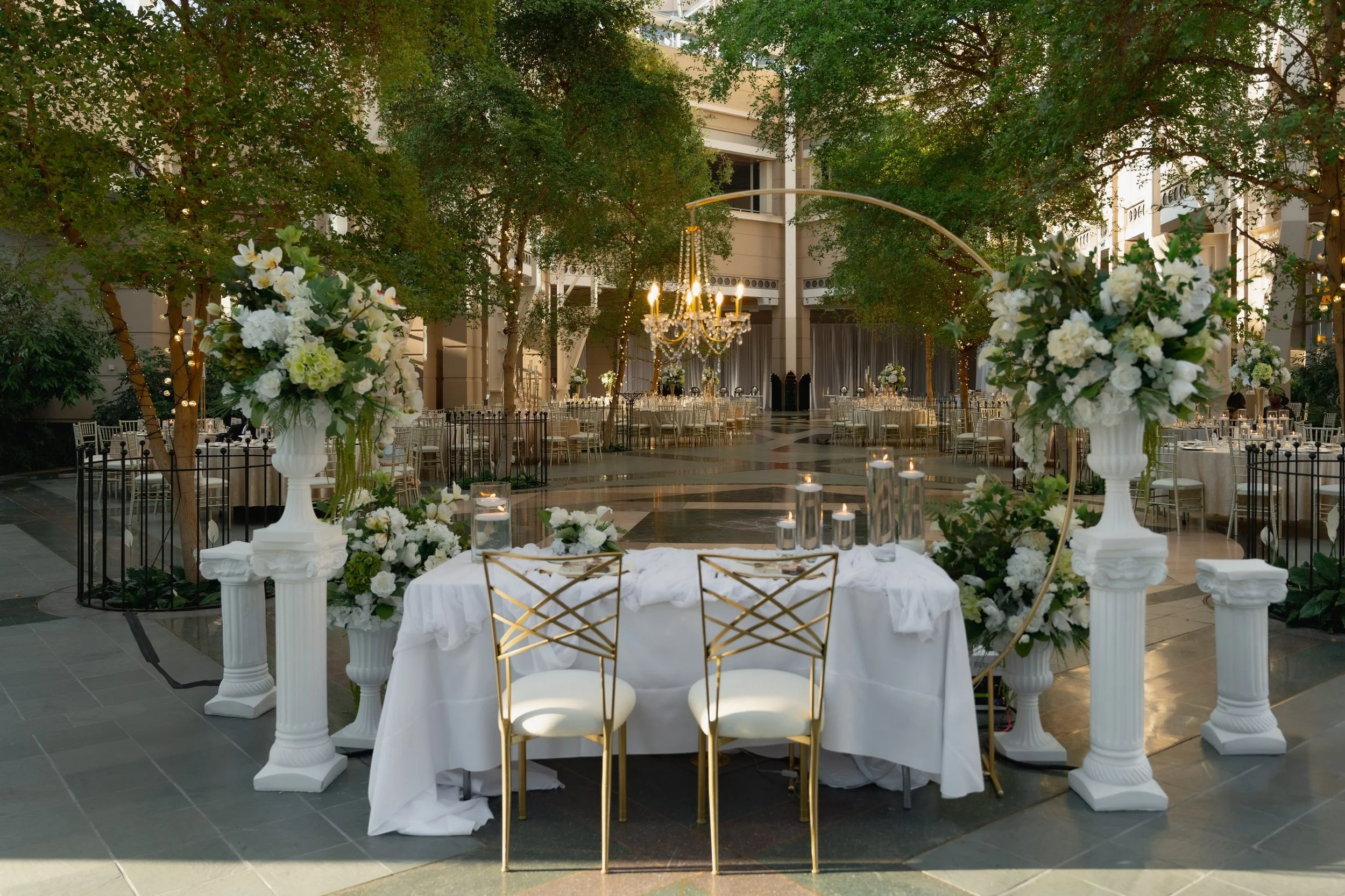 Elegant indoor venue decorated with white flowers and candles for a wedding or special event, featuring a chandelier, trees with string lights, and round tables with chairs.