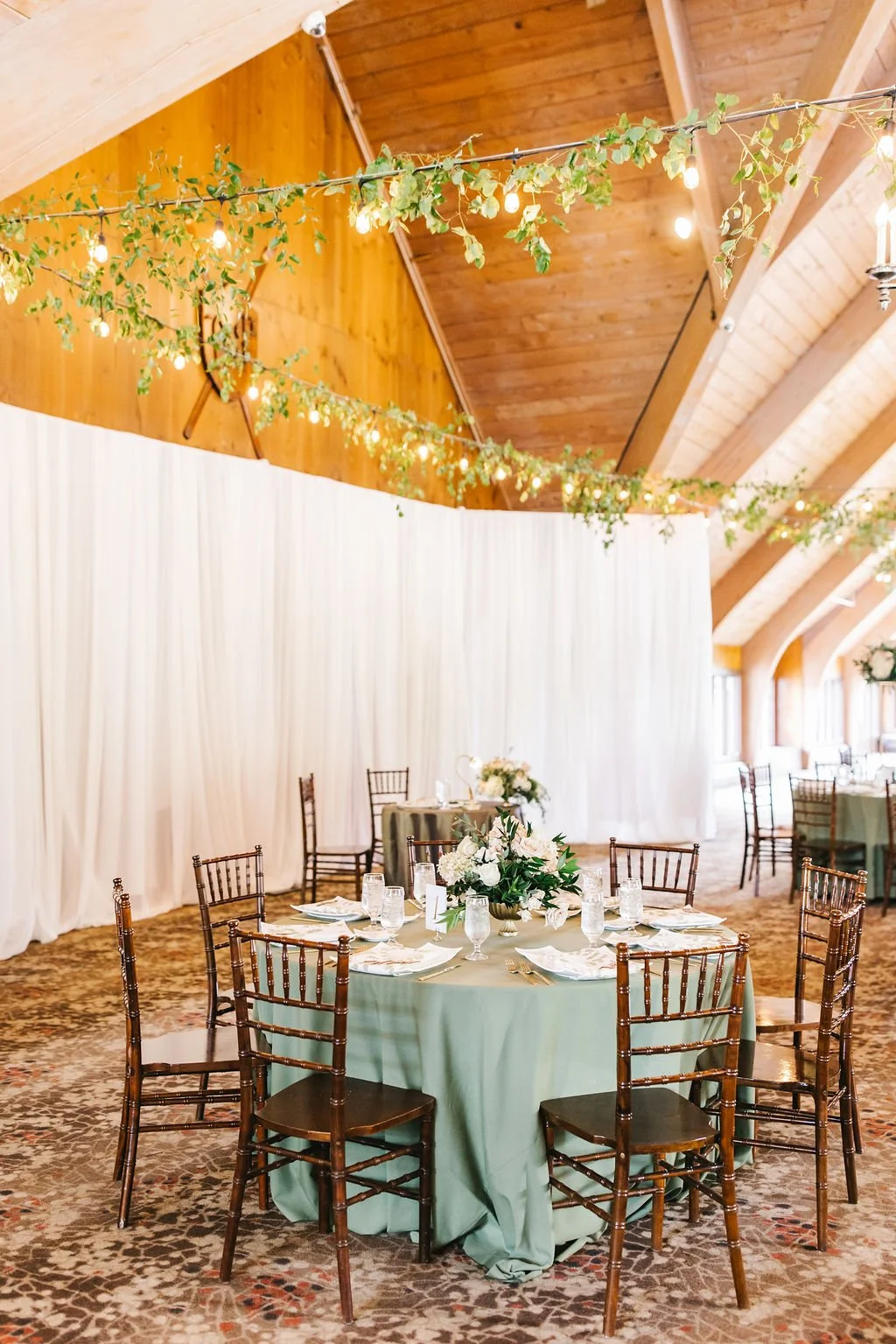 A decorated banquet hall with a round table covered in a sage green tablecloth, set with glassware, plates, and napkins, and adorned with a floral centerpiece. White drapes cover the wall behind the table, and green garlands with string lights hang f