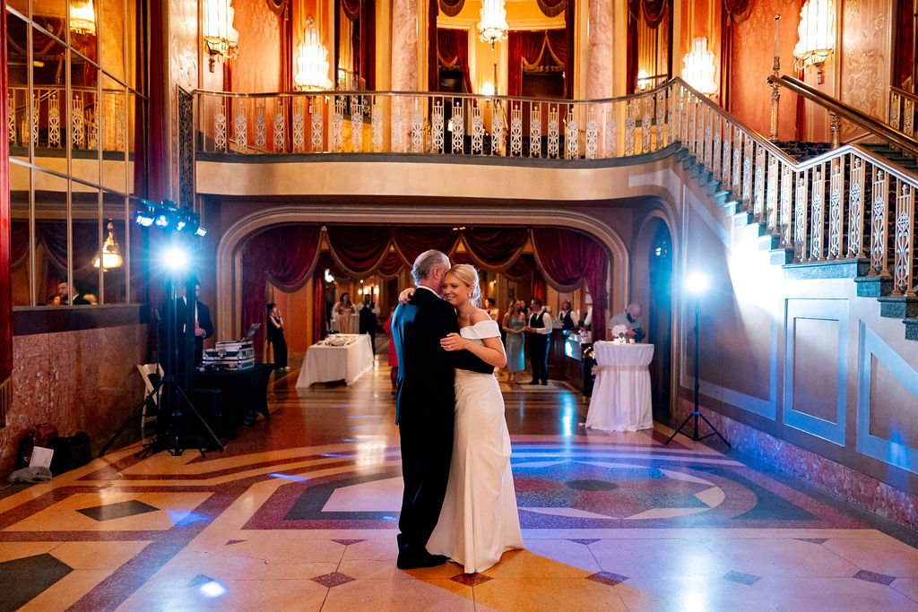 A bride and an older man dancing in a grand ballroom during a wedding reception.