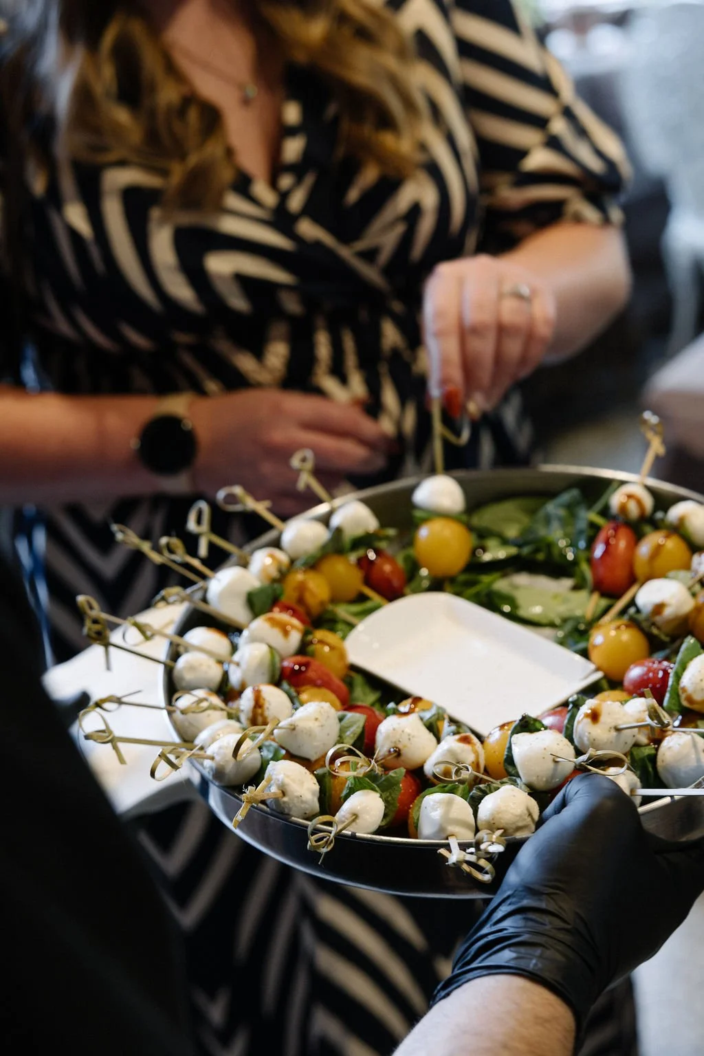 A server holding a round tray of caprese salad bites with skewers, tomatoes, mozzarella, and basil, offering appetizers at an event with a woman in a black and white patterned dress in the background.