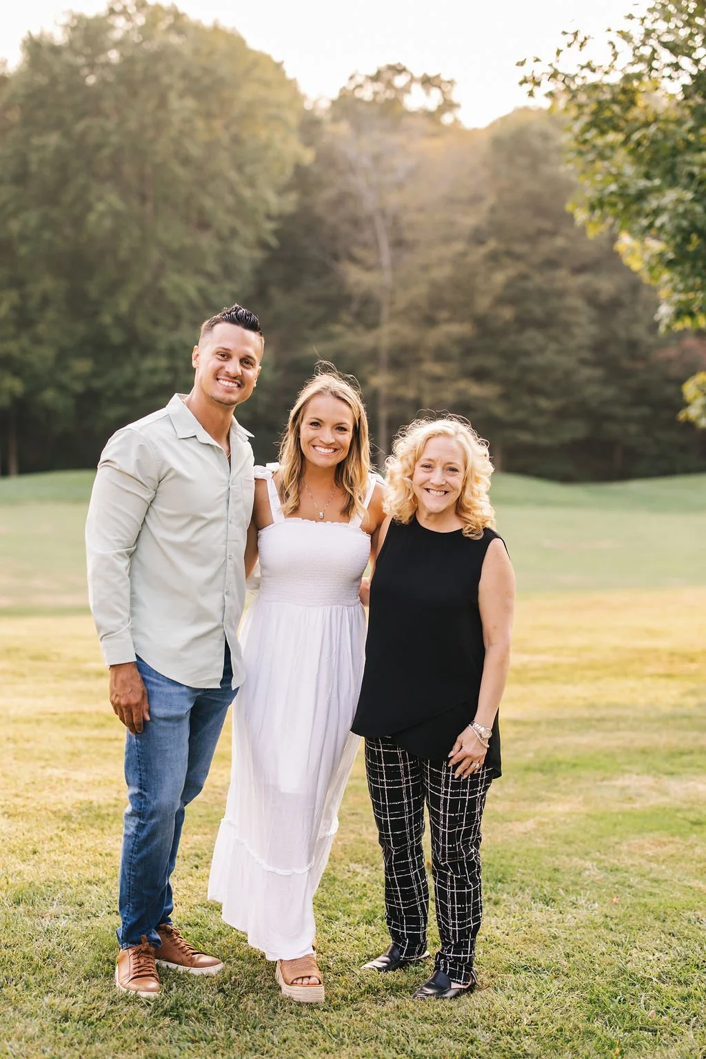 Three smiling people standing outdoors on a grassy field with trees in the background during sunset.