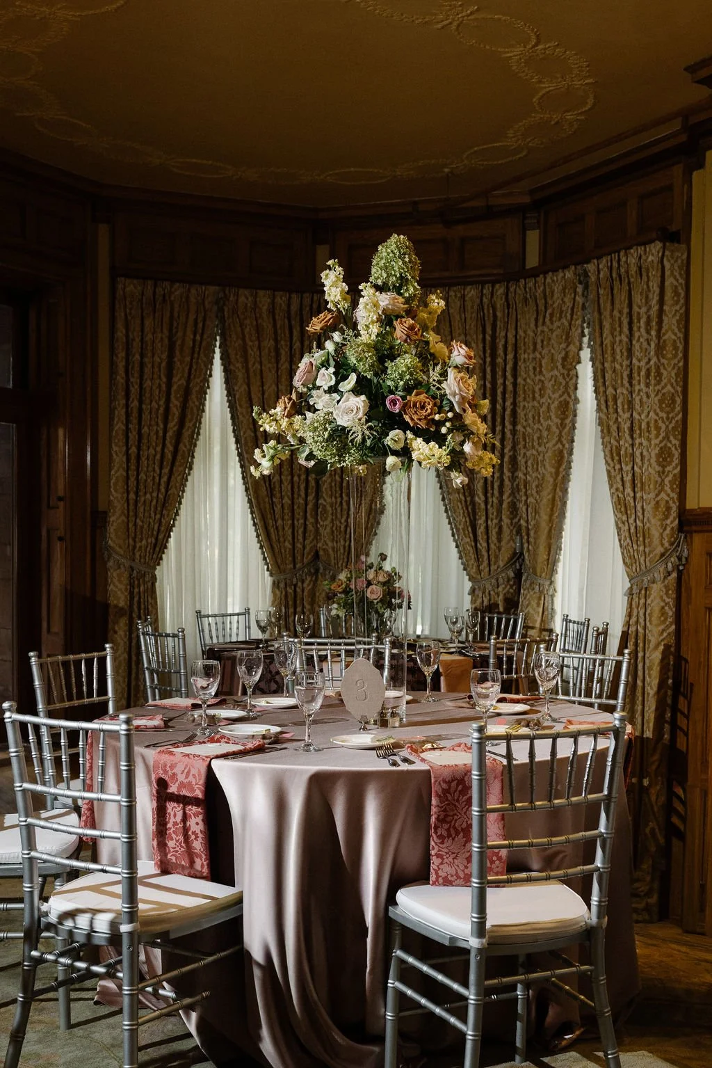Elegant formal dining setup with a round table topped with a luxurious tablecloth, surrounded by silver Chiavari chairs, and decorated with a tall floral centerpiece and glassware.