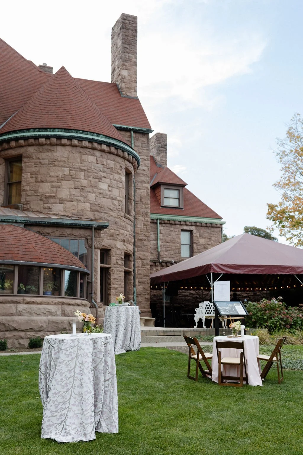 Outdoor event setup featuring round tables with white tablecloths and floral centerpieces, a canopy tent, and a historic stone building with red roofs and chimneys in the background.