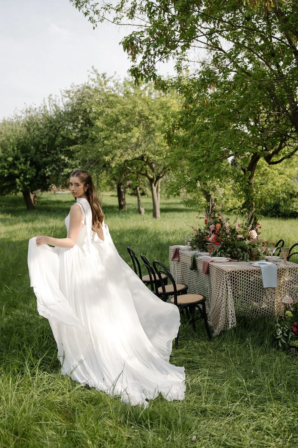 A young woman in an elegant white dress standing in a lush green outdoor setting with trees. There is a table decorated with flowers and tableware nearby, suggesting a garden celebration or wedding event.