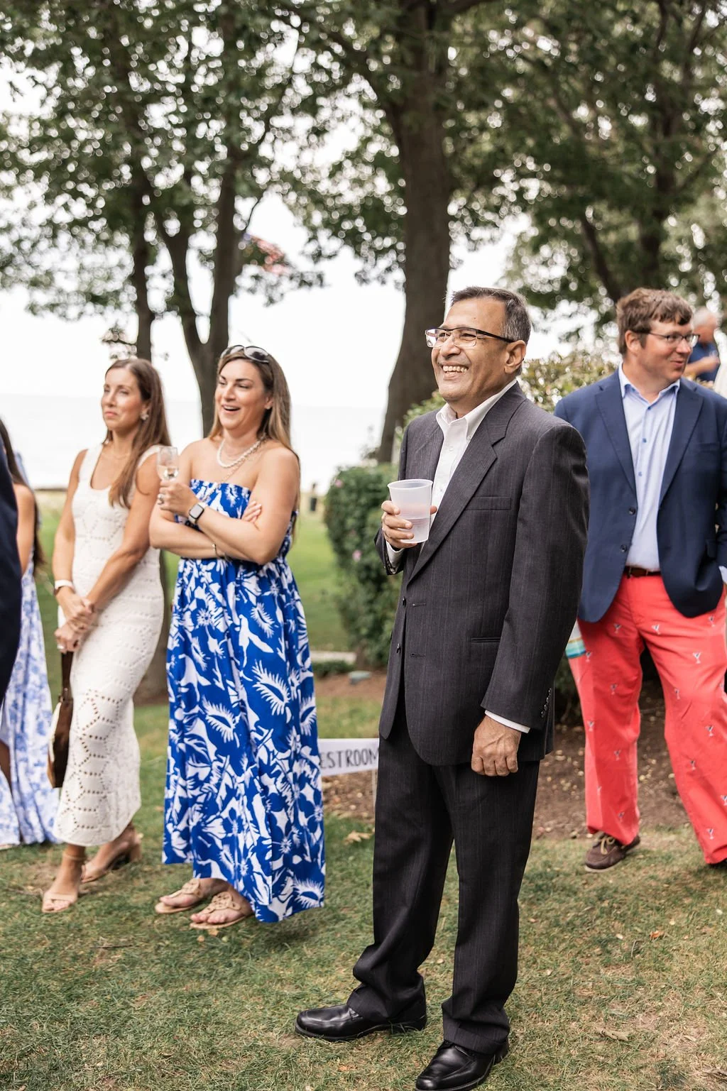 Group of people gathered outdoors, some holding drinks, smiling and socializing among trees and greenery during a daytime event.
