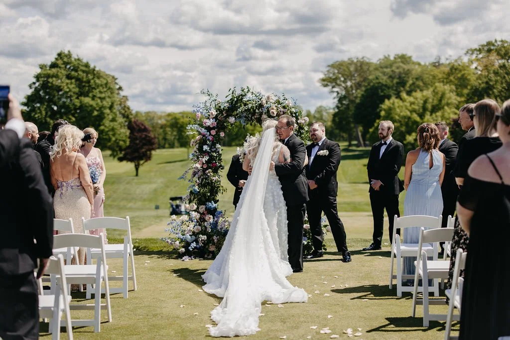Bride and groom kissing under a floral wedding arch on a grassy outdoor field, with guests watching and standing around.