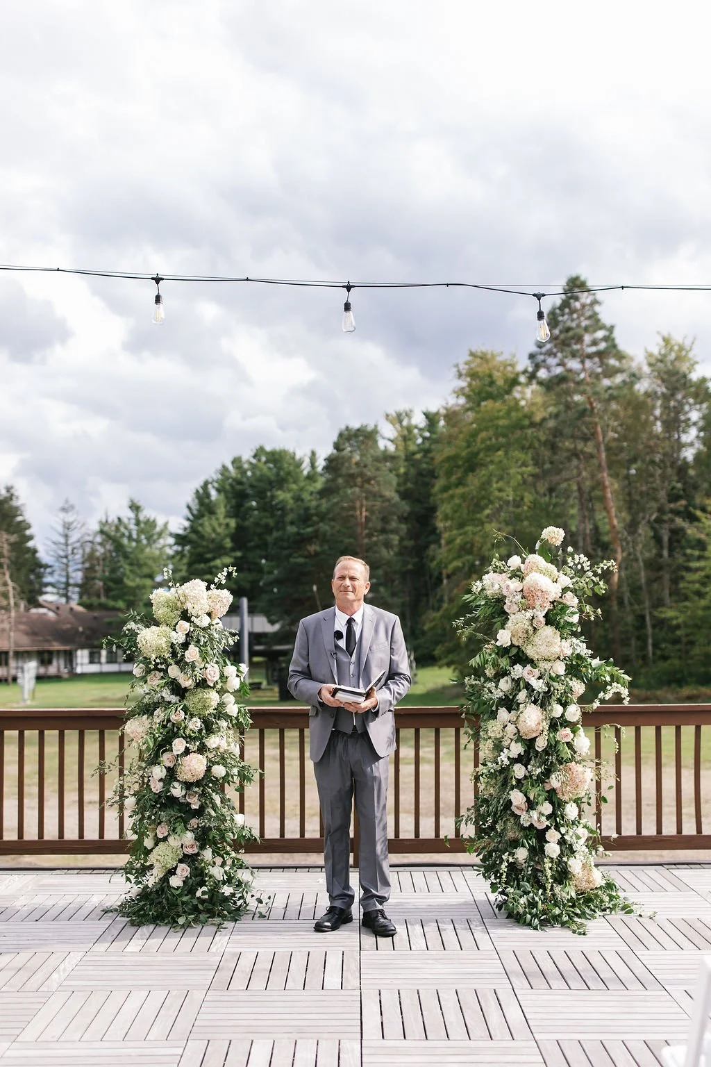 A man in a gray suit standing on a wooden deck with floral arrangements on either side, outdoors during the day with trees and cloudy sky in the background.