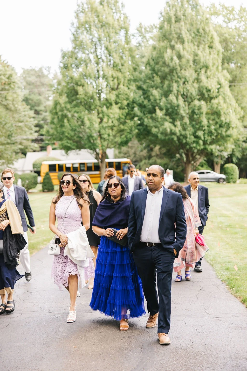 Group of people dressed in formal attire walking outdoors on a paved pathway surrounded by green trees and grass during daytime.