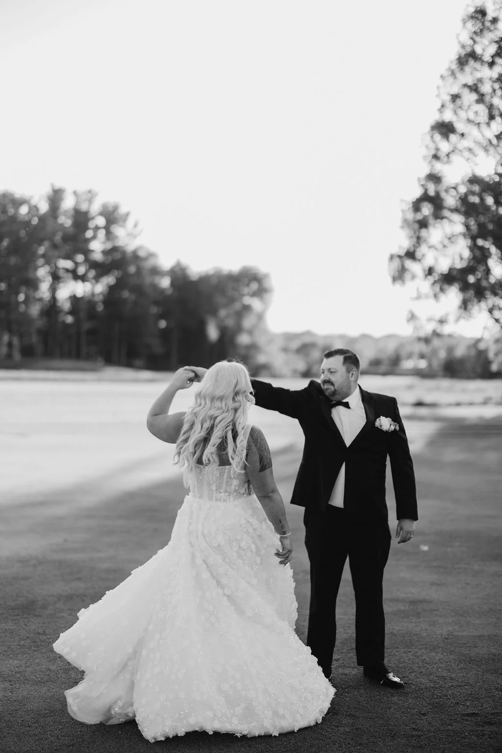Black and white photo of a bride and groom dancing outdoors, the groom with his hand on the bride's head, on a grassy field with trees in the background.