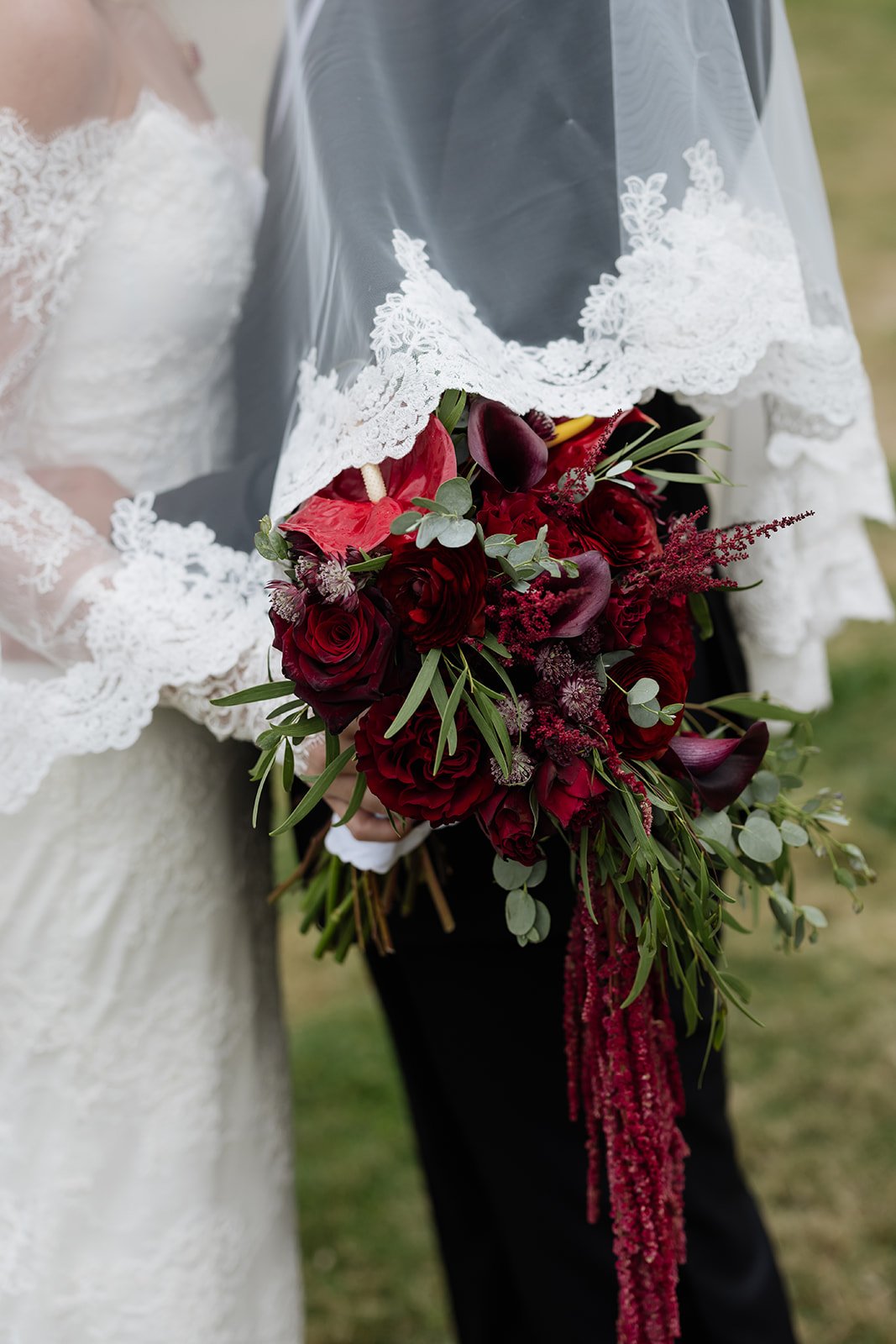 Close-up of a bride holding a cascading bouquet of deep red and burgundy flowers with green foliage, partially covered by a lace-trimmed veil, during a wedding.