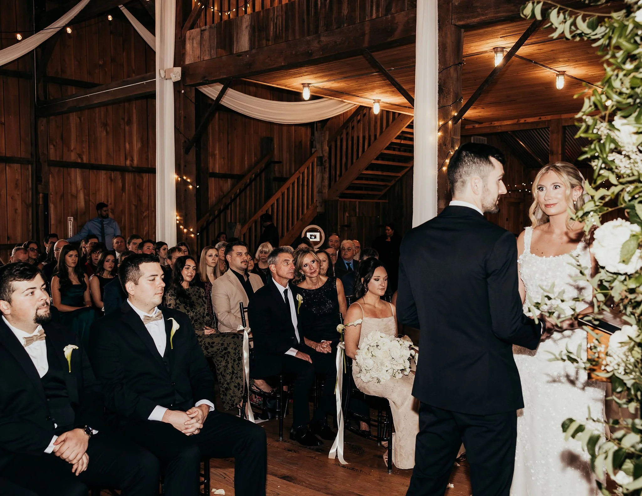 A wedding ceremony inside a rustic wooden venue with string lights and drapery. The bride and groom stand facing each other, with the bride holding a bouquet of white roses. The guests are seated and attentively watching the couple.