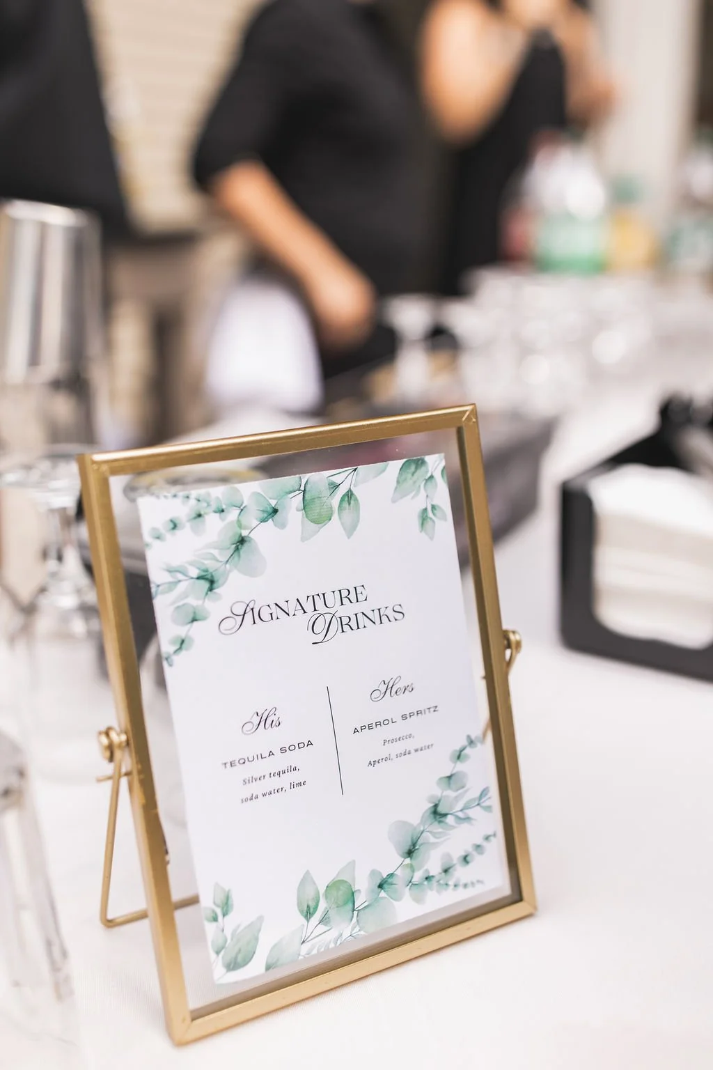 A framed menu titled "Signature Drinks" with a green leafy design, listing two drinks: a His drink with tequila soda, and a Her drink with Aperol spritz, on a white table at an event. Blurred people and serving items are in the background.