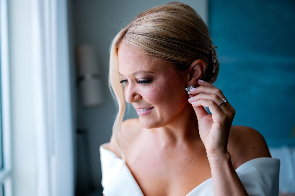 A woman in a white dress putting on diamond earrings in a well-lit room.