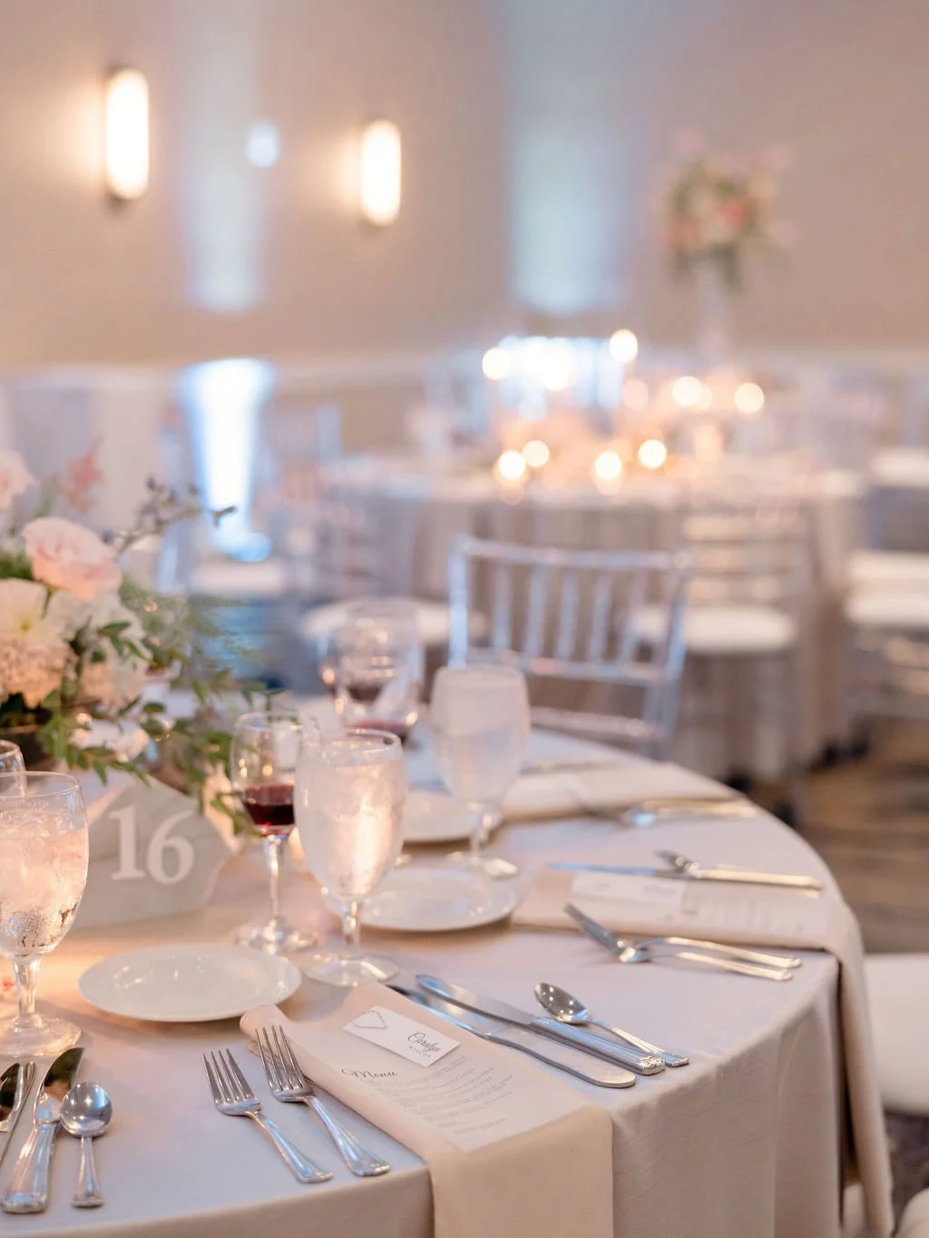Elegant banquet table set with white tablecloth, glassware, silverware, menus, floral centerpiece, and numbered place card in a softly lit reception hall.