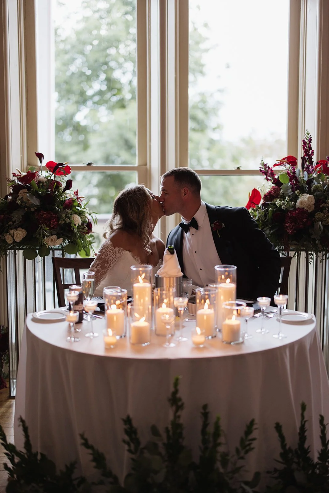 A bride and groom sharing a kiss at their wedding reception, sitting at a decorated table with candles, surrounded by floral arrangements and large windows with trees outside.