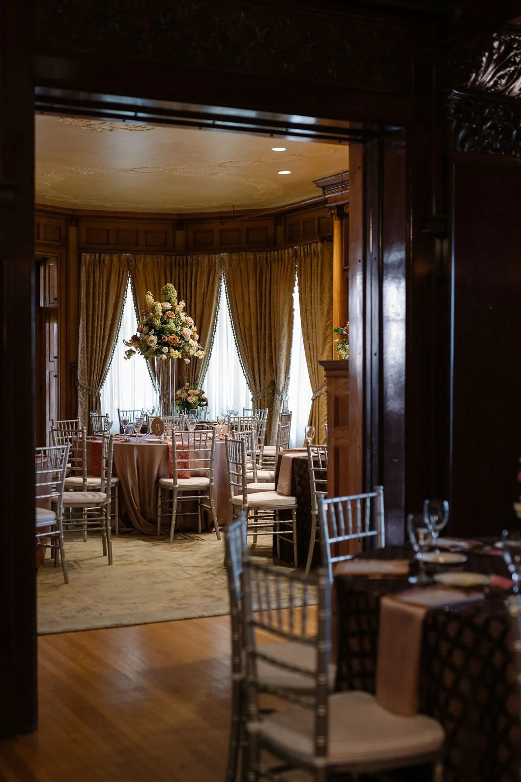 Elegant banquet room with round tables decorated with pink tablecloths and floral centerpieces, gold curtains, and natural light filtering through windows.