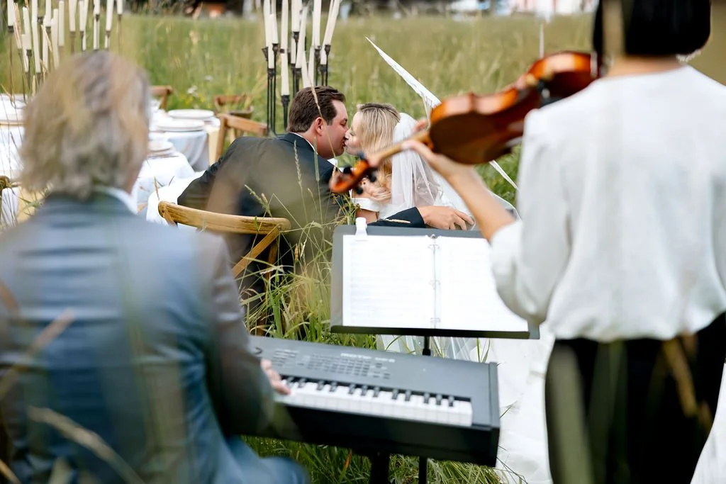 A bride and groom sharing a kiss at an outdoor wedding ceremony, with musicians playing instruments nearby.