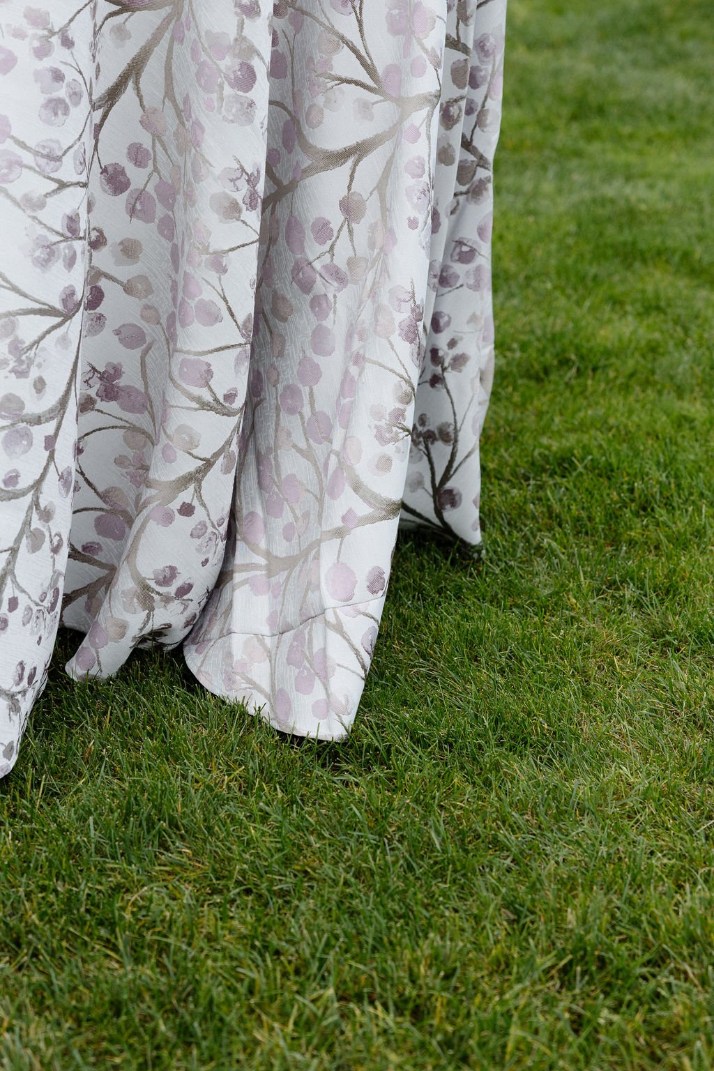 A fabric curtain with a floral pattern of purple and beige branches and berries, hanging outdoors on green grass.