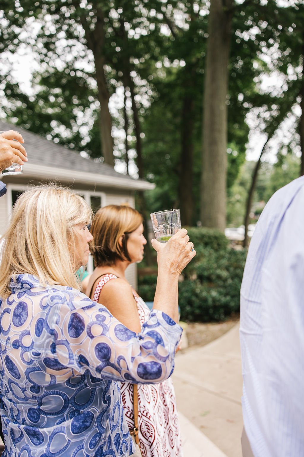People gathered outdoors, holding glasses with drinks, under tall trees.