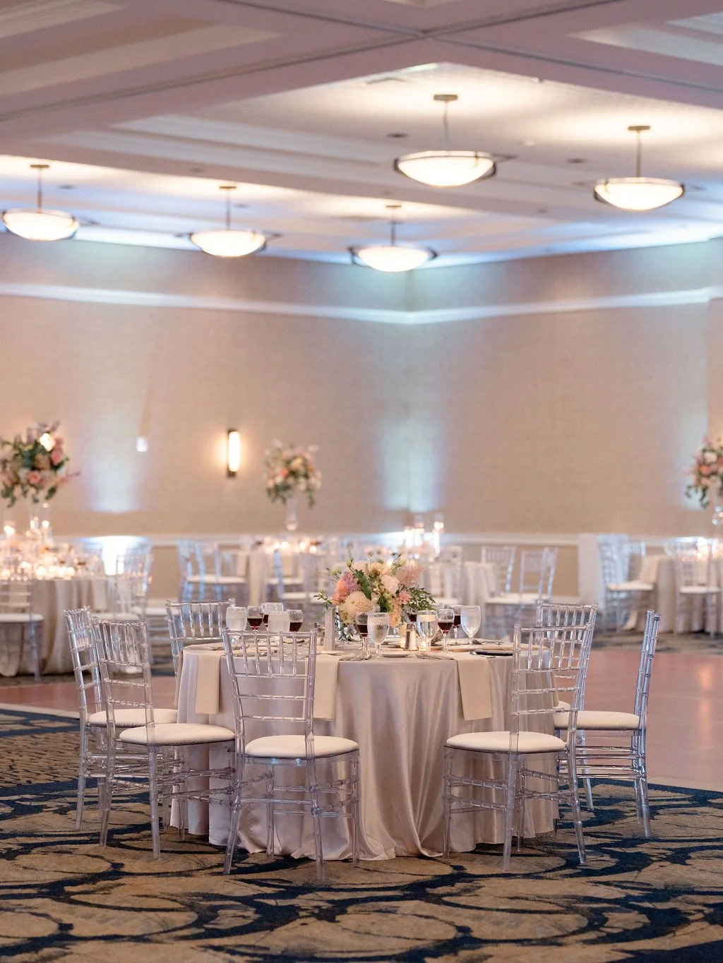 Elegant banquet hall decorated with floral centerpieces, round tables with white tablecloths, and crystal chairs, set for a celebration or wedding reception.