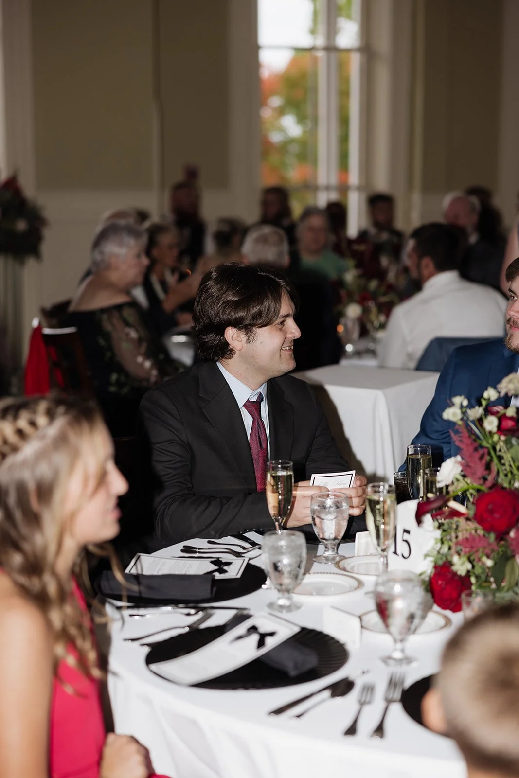 A man in a black suit with a red tie sitting at a formal event table, smiling and holding a card, surrounded by other guests and floral centerpieces.