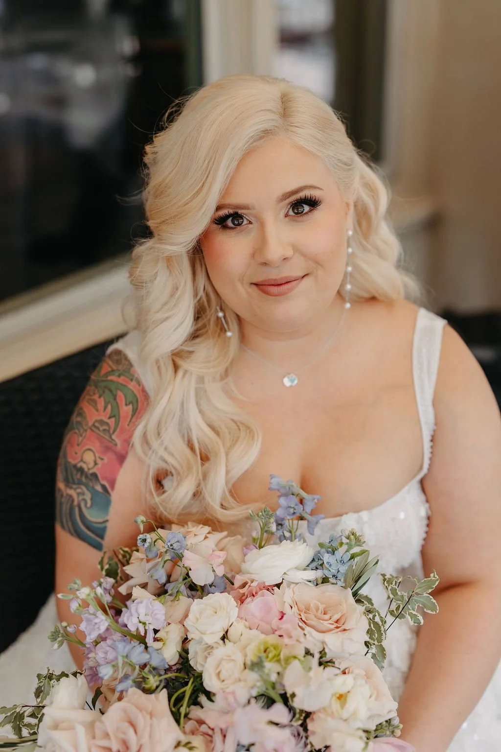 A smiling woman with long blonde hair, wearing a white dress, holding a bouquet of pastel-colored flowers, and sitting indoors.