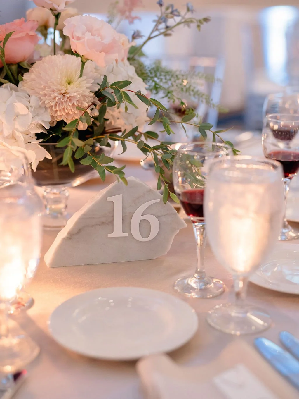 A table centerpiece with pink and white flowers and green foliage, a white stone table number 16, and wine glasses filled with red wine, set for an event.