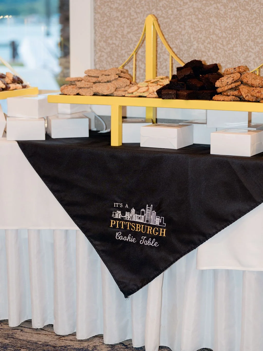 A dessert table with stacks of various cookies on a yellow trellis display, underneath a black tablecloth with 'It's a Pittsburgh Cookie Table' embroidery, set in an indoor event space.