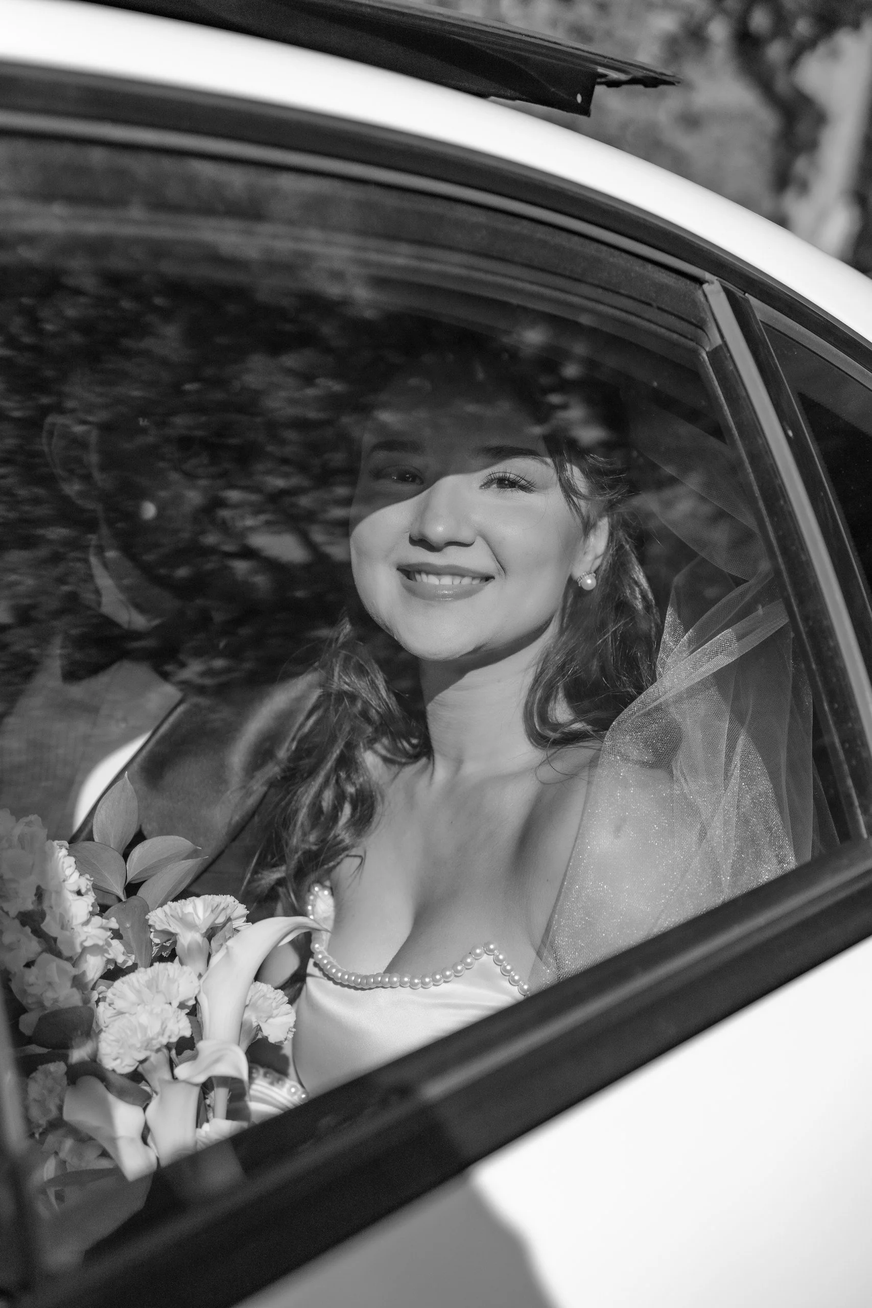 A black and white photo of a woman sitting inside a car, smiling and looking out the window. She is wearing a pearl necklace and earrings, with curly hair and a wedding dress, holding a bouquet of flowers. The car's window reflects trees outside.