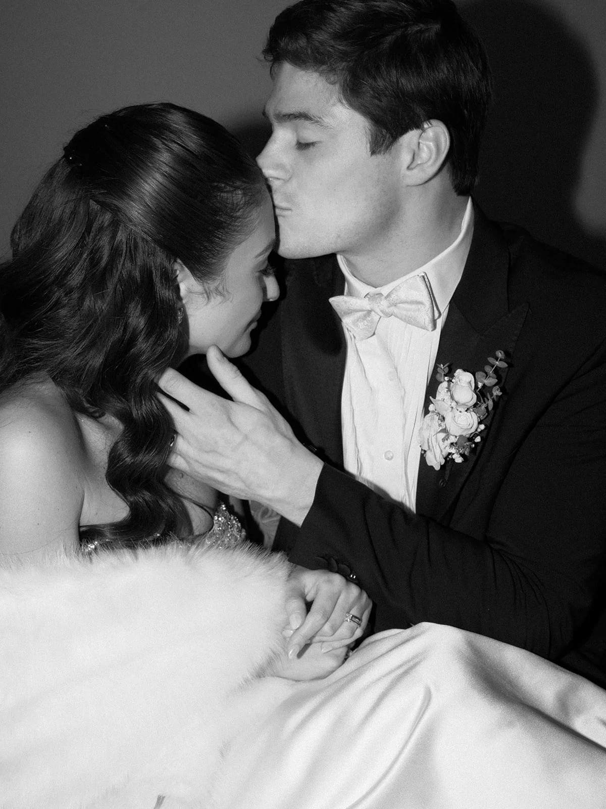 A black and white photo of a bride and groom embracing during their wedding. The groom is kissing the bride on her forehead, and the bride has her eyes closed with her hand resting on the groom's chest. The bride is wearing a strapless wedding dress 
