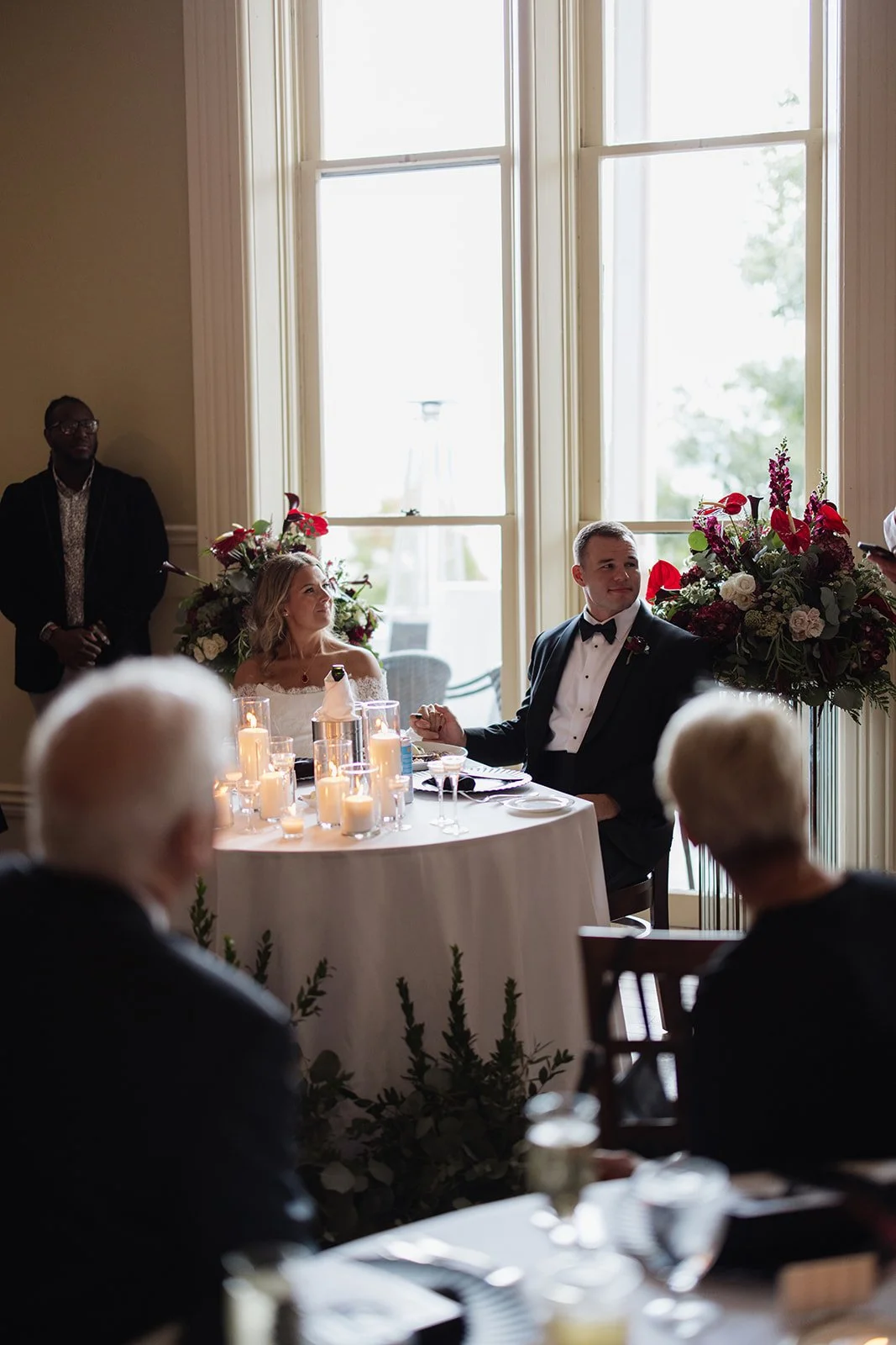 A wedding reception with a bride and groom sitting at a table with candle centerpieces, flowers, and glassware while guests watch, with a large window in the background.
