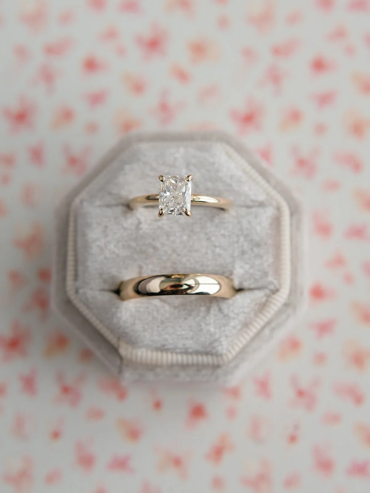 A jewelry box with two gold rings, one with a large rectangular diamond, on a pink and white patterned surface.