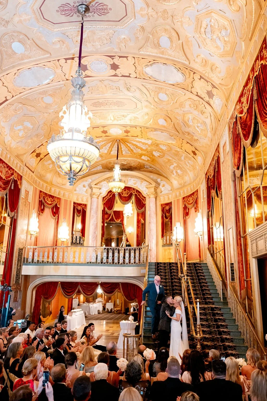 A wedding ceremony in an elegant, ornate hall with a high, decorated ceiling and red drapes. The bride and groom are kissing on the staircase in front of an officiant, while guests seated and standing around them applaud and take photos.