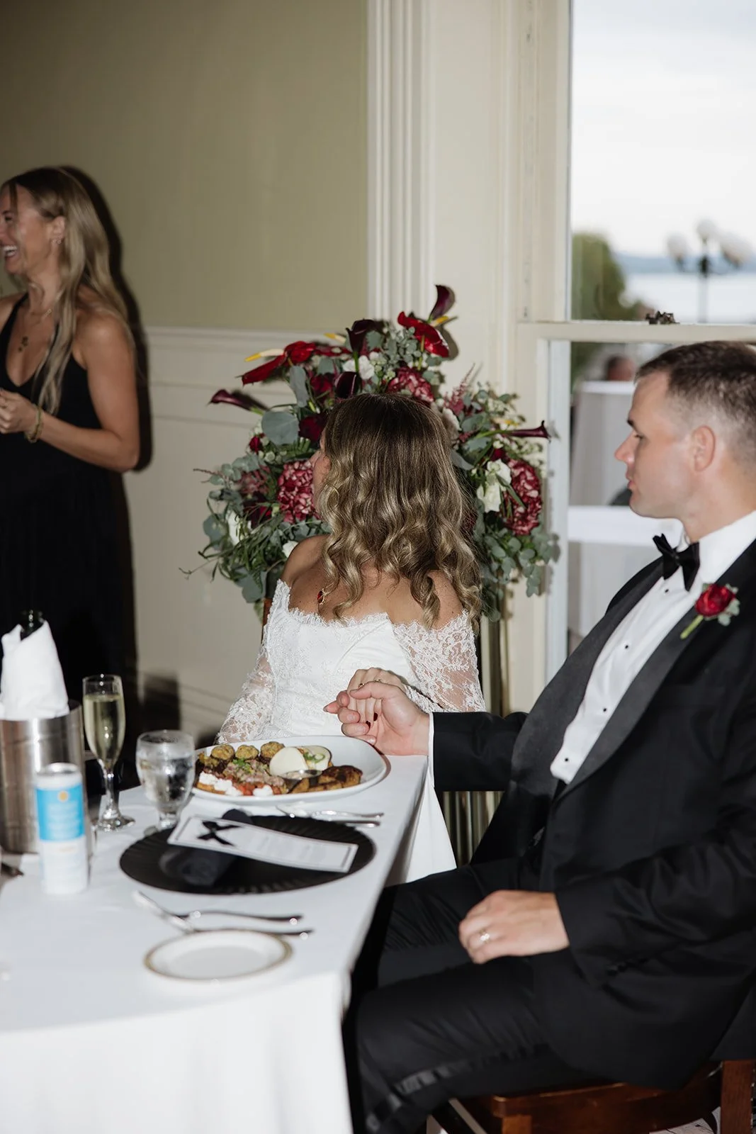 A wedding reception scene with a bride and groom seated at a table, decorated with a large floral arrangement, with a woman standing and smiling in the background.