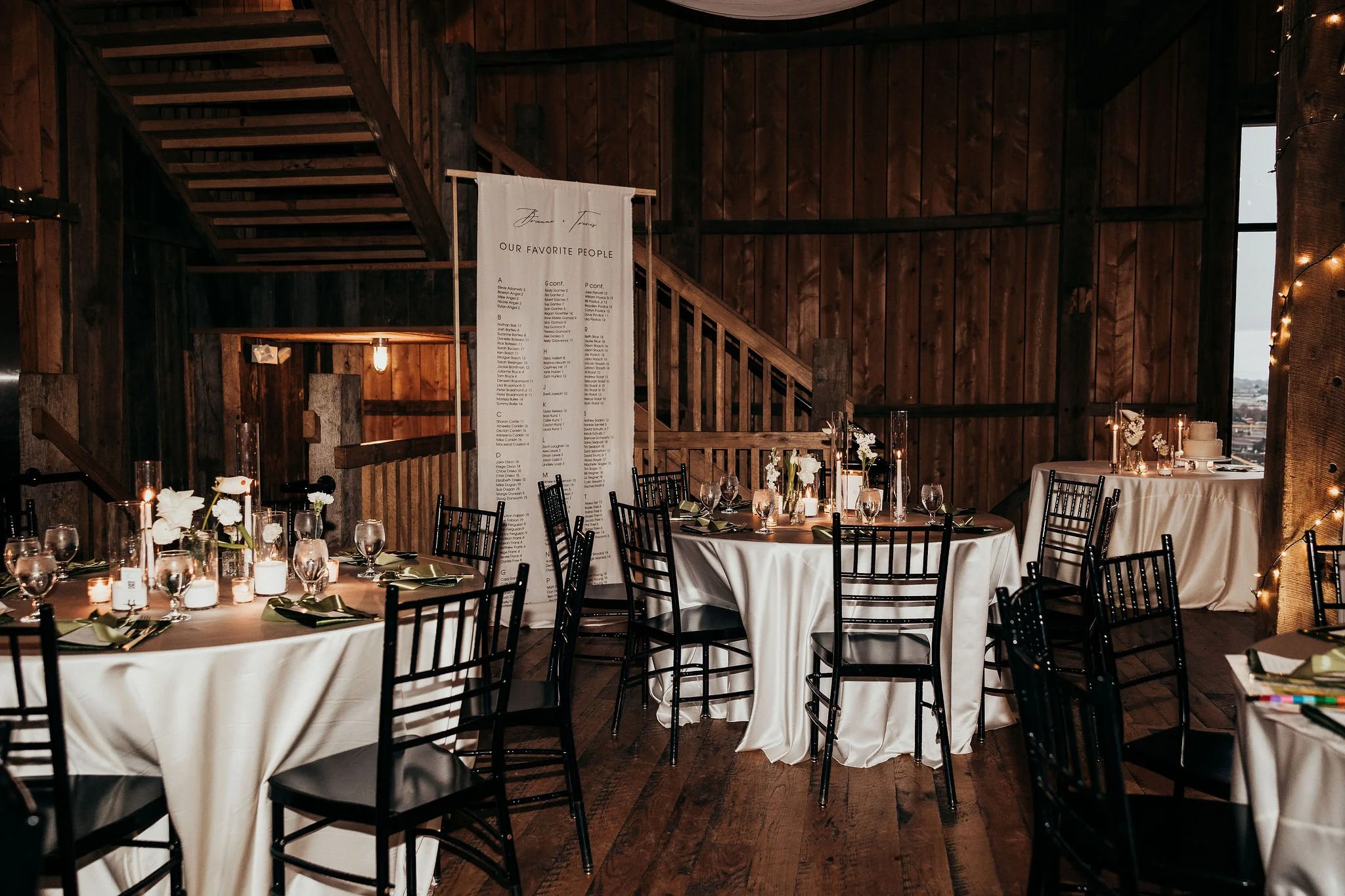 Indoor wooden venue decorated for a celebration with round tables covered in white tablecloths, black chairs, candles, and floral arrangements. A large white sign lists names with the heading 'Our Favorite People,' and string lights are draped on the