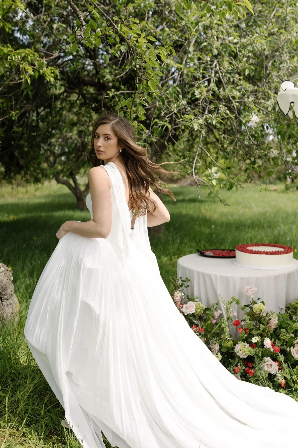 Young woman in a white dress standing outdoors with trees and green grass, beside a table with a floral arrangement and a cake with red berries.